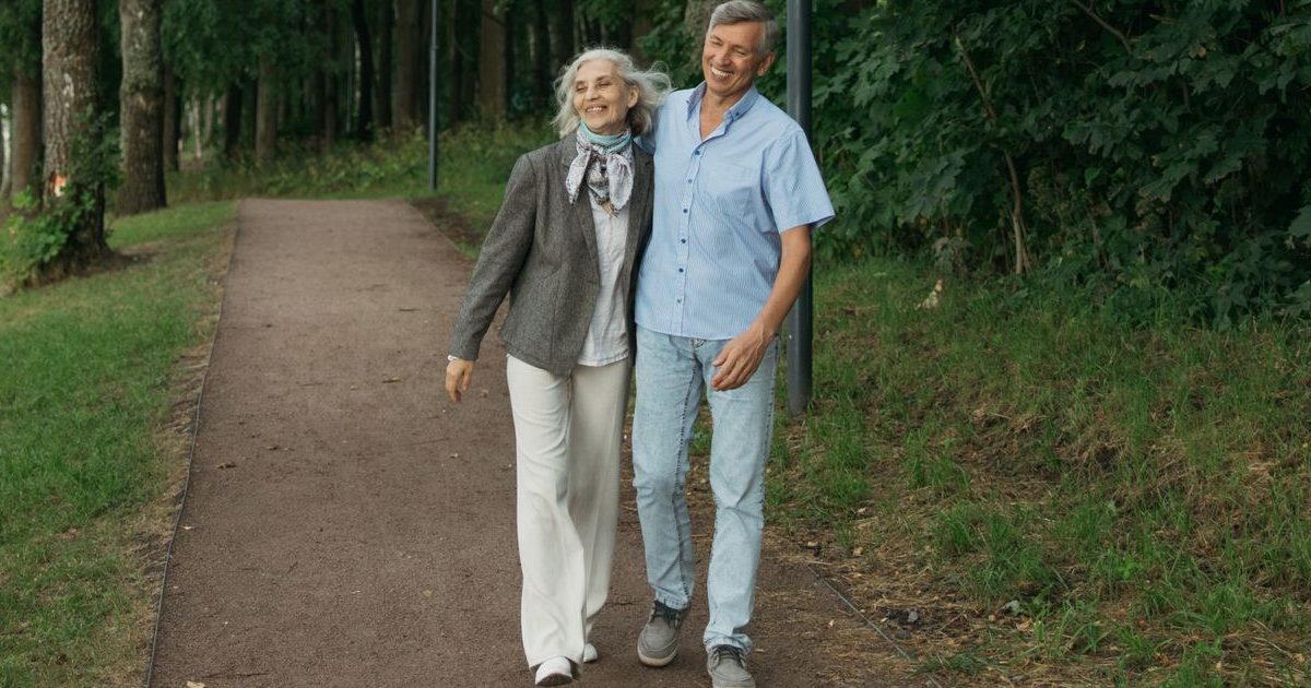 A smiling older couple walk arm in arm along a tree lined park path on a summer day