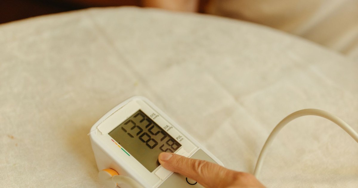 A white home blood-pressure monitor on a cream cloth showing a reading in mmHg, with a fingertip pressing its start button and the cuff tube curving to the right