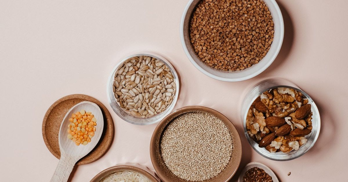 An overhead flatlay of bowls of seeds, nuts, and whole grains on a soft neutral surface