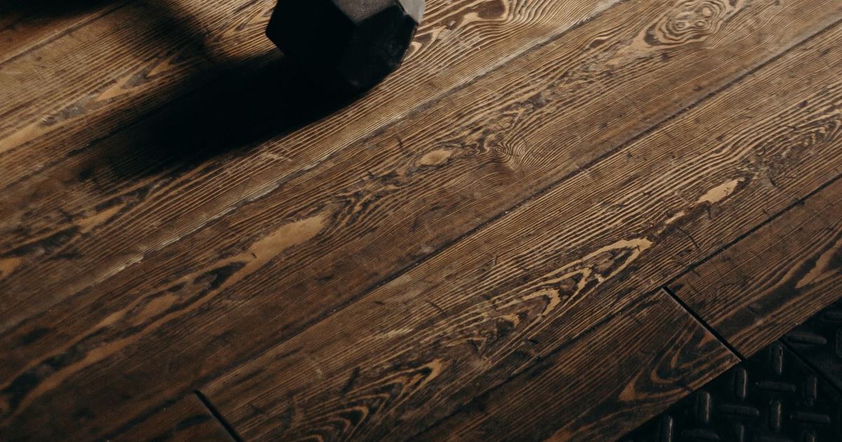 A single black hex dumbbell on a warm dark-wood floor with a long side-lit shadow